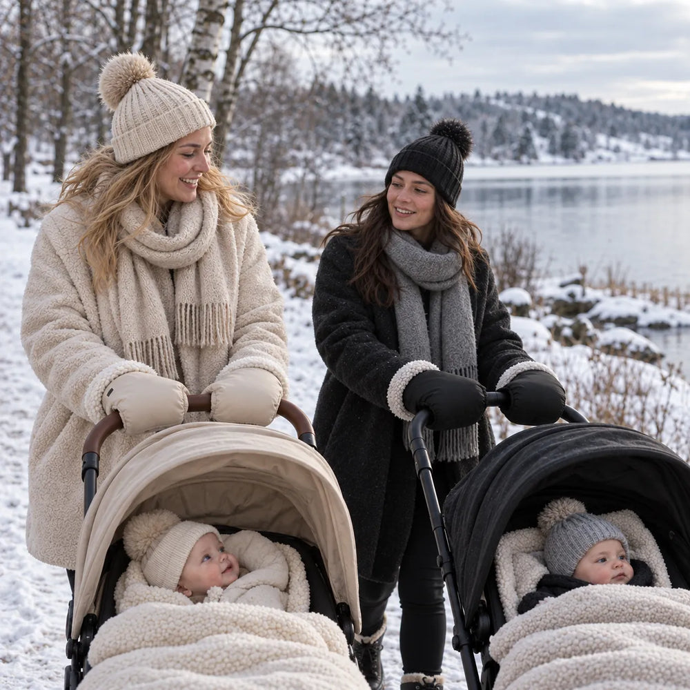 Twee moeders wandelen met kinderwagens in de sneeuw – de een met beige en de ander met zwarte Lovelle Cosy handwarmers